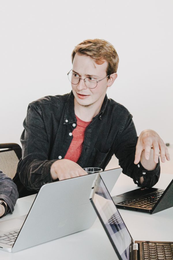 Portrait of an employee explaining something, seated at a table with three laptops.